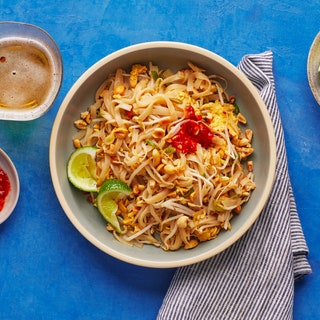 Bowl of pad Thai rice noodles topped with bean sprouts limes sambal oelek and peanuts with a glass of beer on the side.